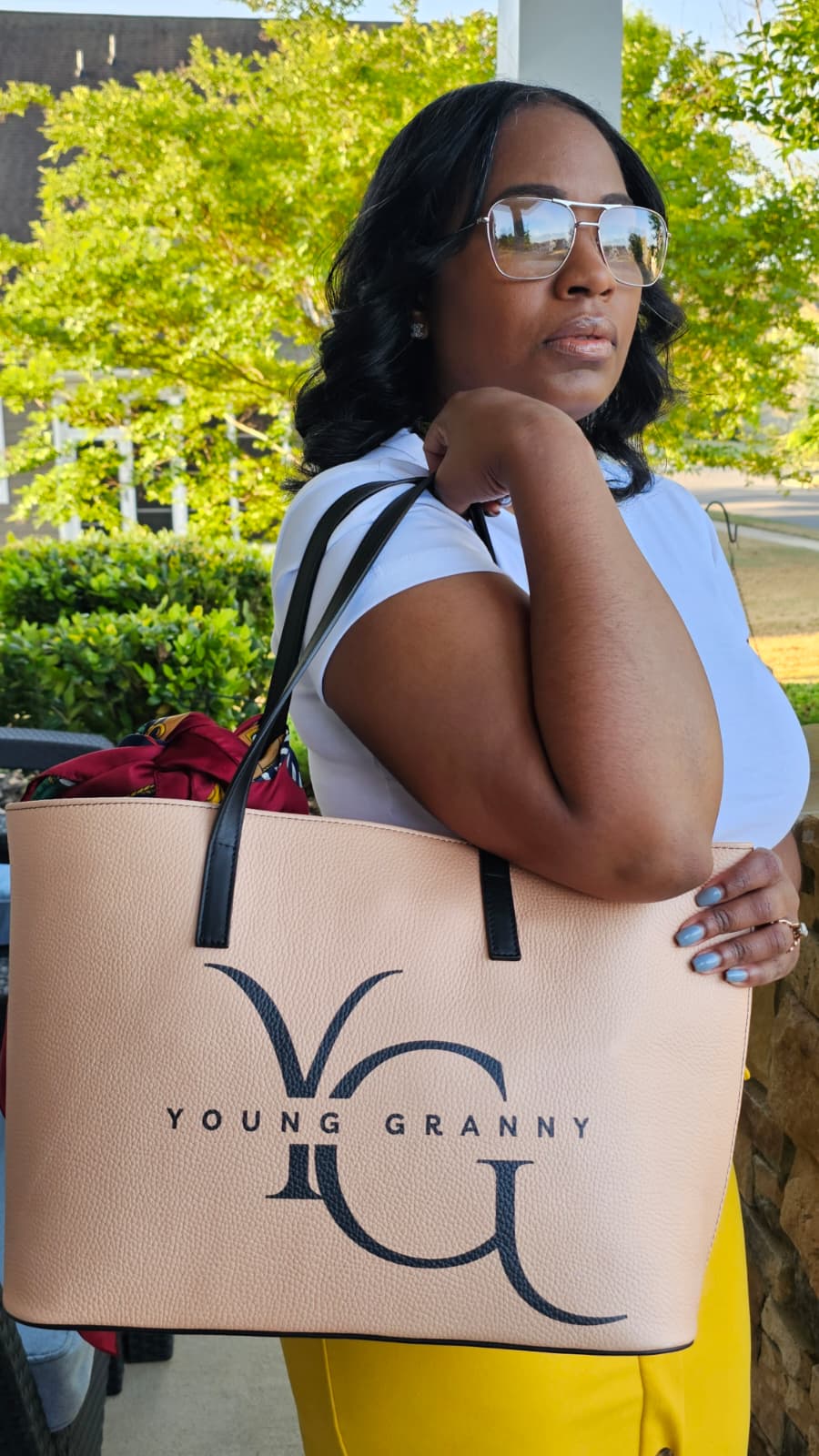 Woman holding a beige small tote bag with 'Young Granny' logo outdoors.